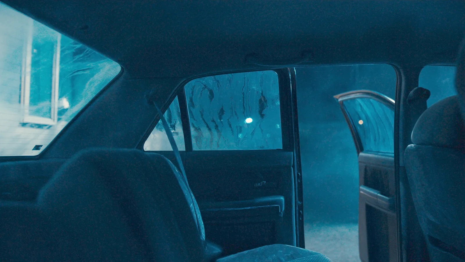 An empty car back seat at night with a door open and city light on the window