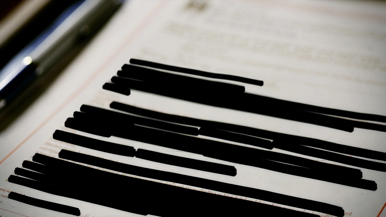 A legal document with heavy redaction bars across it, resting on a desk in raking light