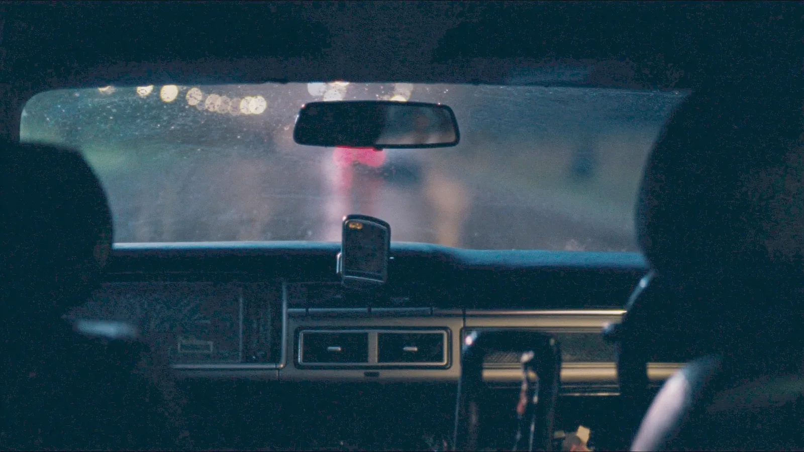 The view forward from a car back seat at night through a rain-streaked windscreen onto an empty street
